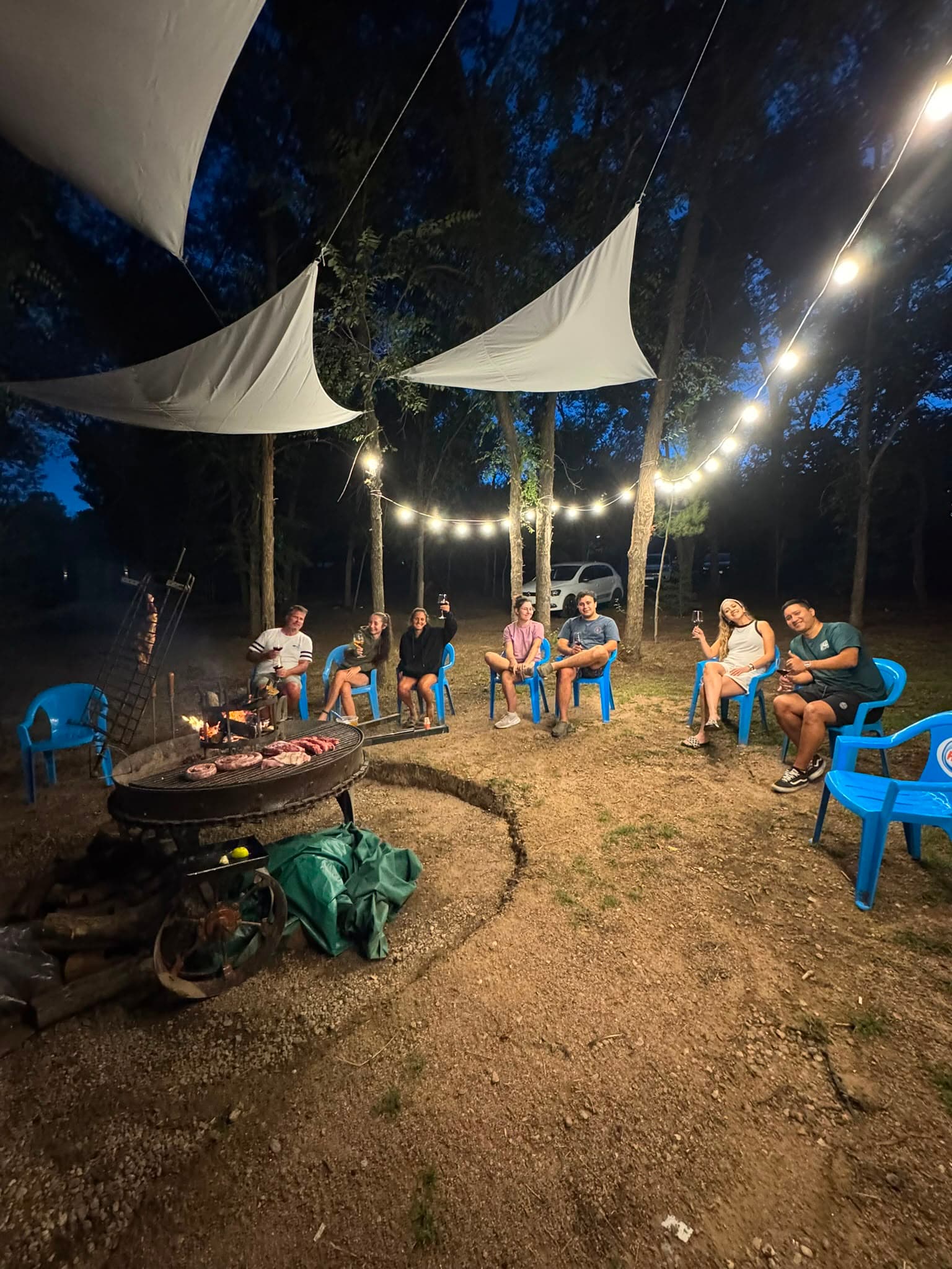 Friends gathered around the asado grill at night, string lights overhead