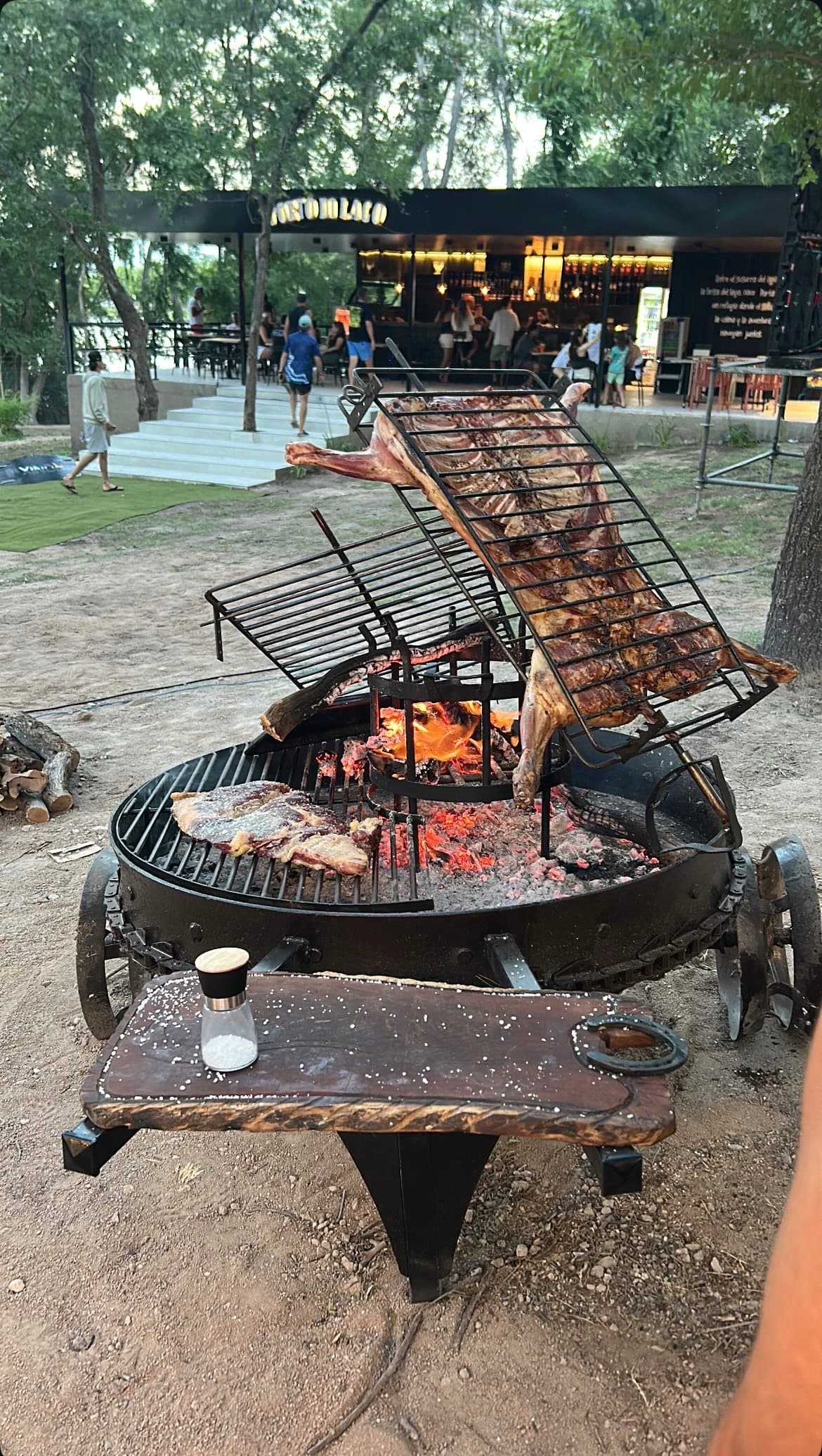 Whole cuts of meat roasting over an open-fire asado grill, Porto di Lago in the background